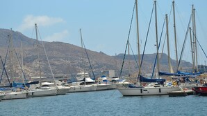 A marina with multiple sailboats docked, a mountainous backdrop, and calm water.
