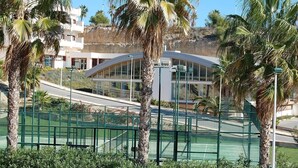 A modern building with a curved glass facade, surrounded by palm trees and a tennis court.
