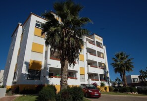 A multi-story residential building with balconies, a car parked in front, and palm trees.