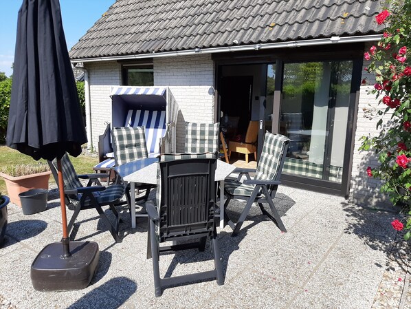 Eine Terrasse im Freien mit Tisch und Stühlen, einem schwarzen Sonnenschirm und einem Gebäude mit einem Ziegeldach und großen Schiebeglasfenstern.