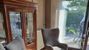 Ein modernes Wohnzimmer mit zwei grauen Sesseln und einem kleinen Hocker, einem Holzschrank mit Glastüren sowie einem Blick auf einen Garten durch eine große Schiebetür.