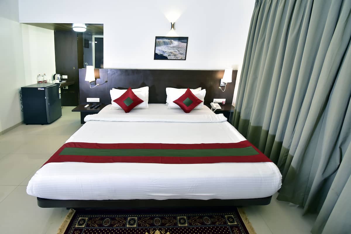 Quarto de hotel com uma cama grande, mesa de cabeceira com luminária, mini-frigorífico, escrivaninha com cadeira e um quadro na parede.