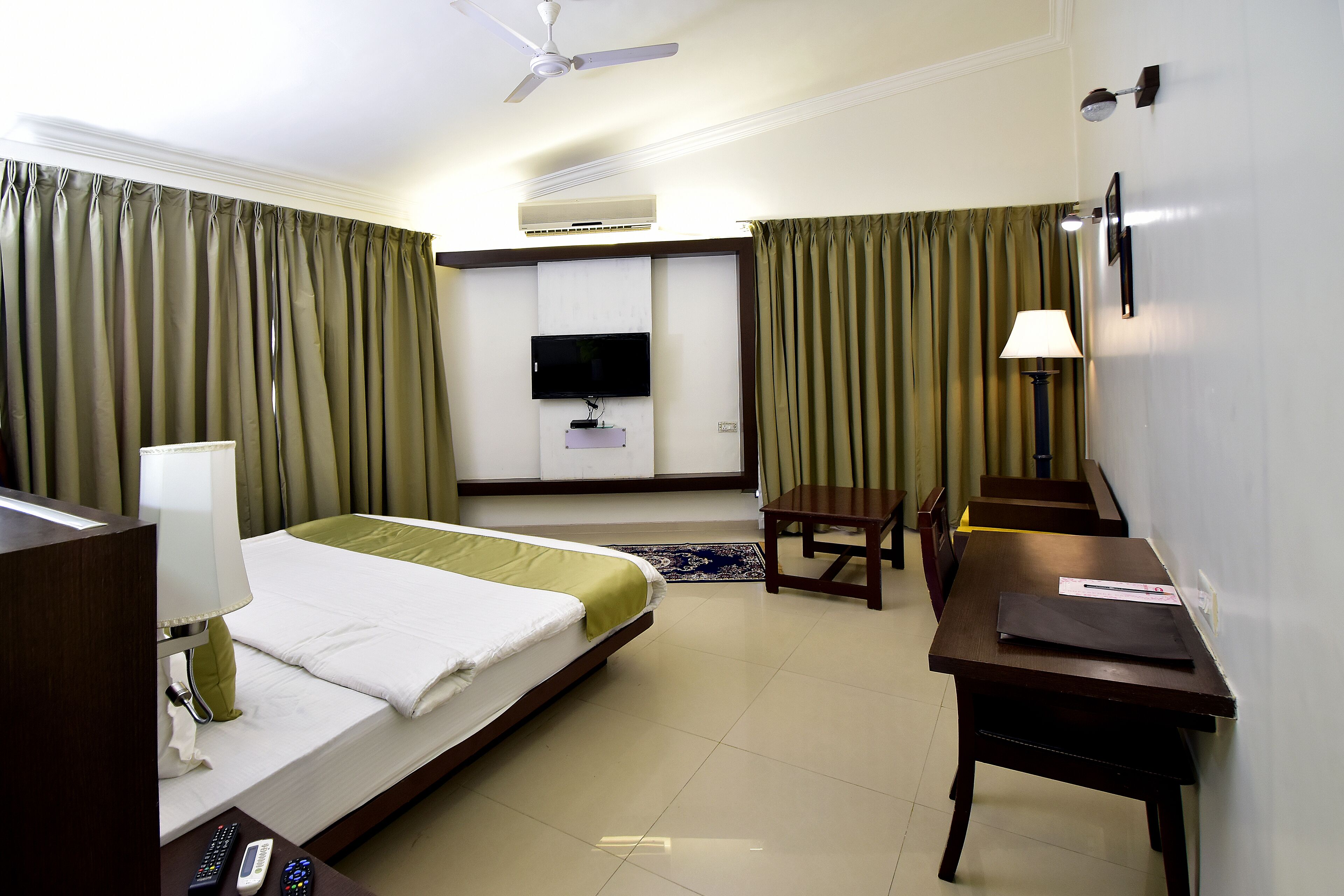 Una habitación de hotel con cama, escritorio, silla, televisión, lámpara y ventilador de techo.
