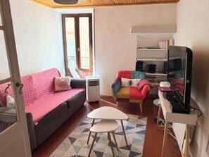 Ein gemütliches Zimmer mit einer Couch, einem bunten Sessel, einem kleinen Tisch und einer TV-Konsole mit Lautsprechern.