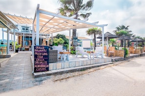 An outdoor seating area with a pergola, lounge chairs, and a signboard advertising grilled kalamari, fresh local fish, Greek specialities, and Mediterranean cuisine.