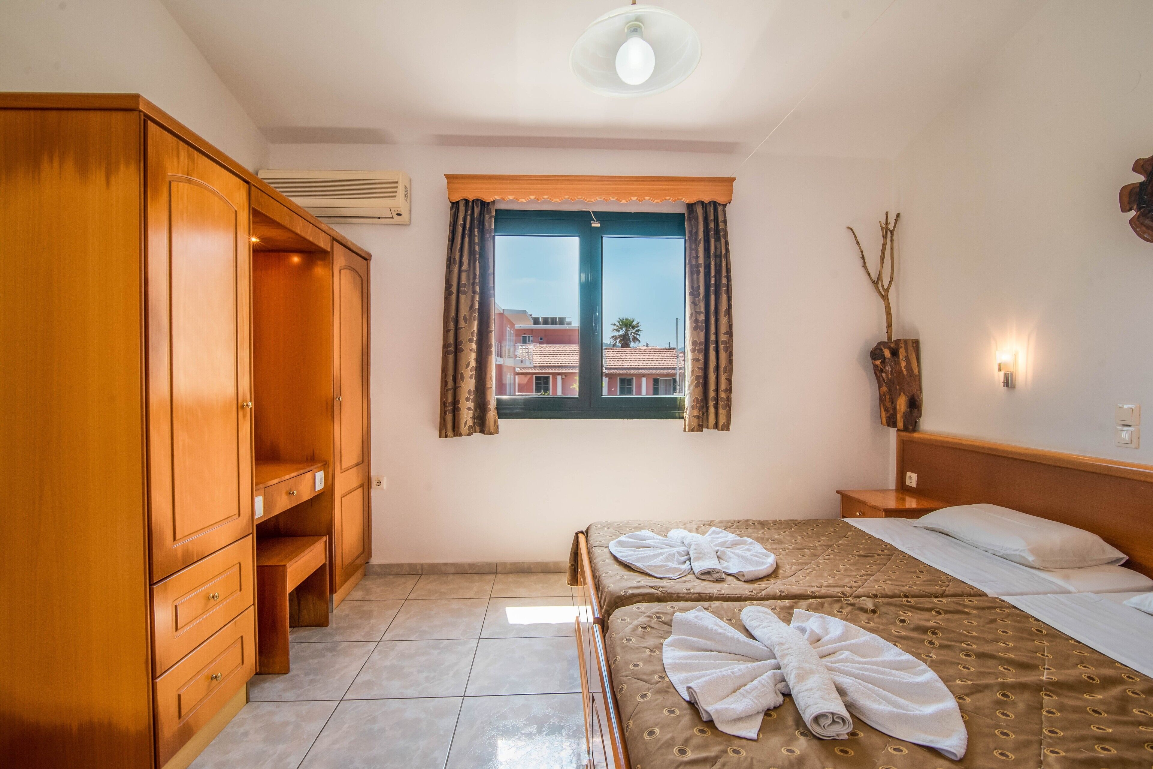 Habitación de hotel con cama, armario, ventana con cortinas, mesita de noche y lámpara de pared.