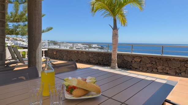 A table with a bottle and glasses, a plate with food, a palm tree, and a view of the sea. - Vacation Rental 'Villa Loretta' with Sea View, Heated Pool & Wi-Fi (Puerto del Carmen)