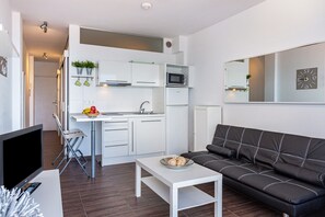 Un salon moderne comprenant un canapé, un téléviseur, une petite table et un coin cuisine avec des placards et un four à micro-ondes.