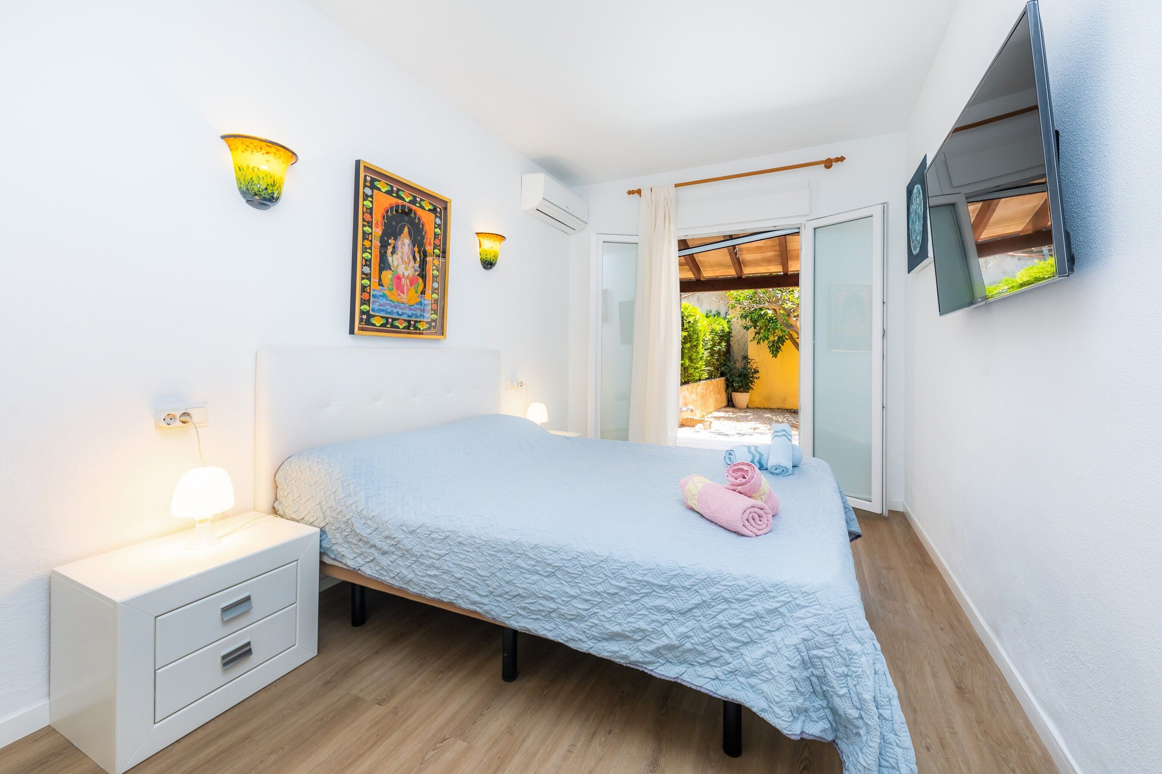 A bedroom with a bed, bedside table, wall-mounted lights, a painting, and a window with a view of greenery.