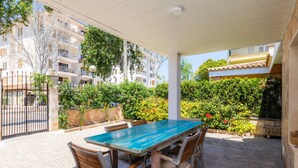 An outdoor patio with a table and chairs, surrounded by greenery and a building in the background.