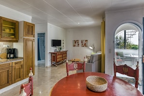 Une cuisine spacieuse avec des meubles en bois, une table à manger avec des chaises, un téléviseur et, à travers une arche, une vue sur un espace extérieur aménagé pour se détendre.
