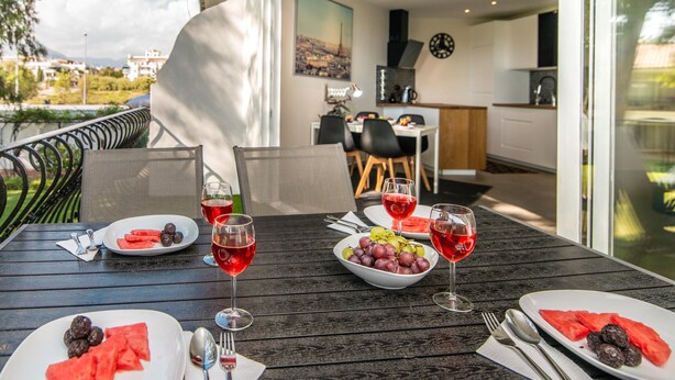 Un área de comedor al aire libre con vistas a la ciudad, una mesa servida con sandía y uvas, y copas de vino tinto.