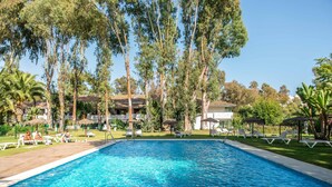 Piscina con sillas de descanso y sombrillas, rodeada de árboles y un edificio con tejado de tejas.