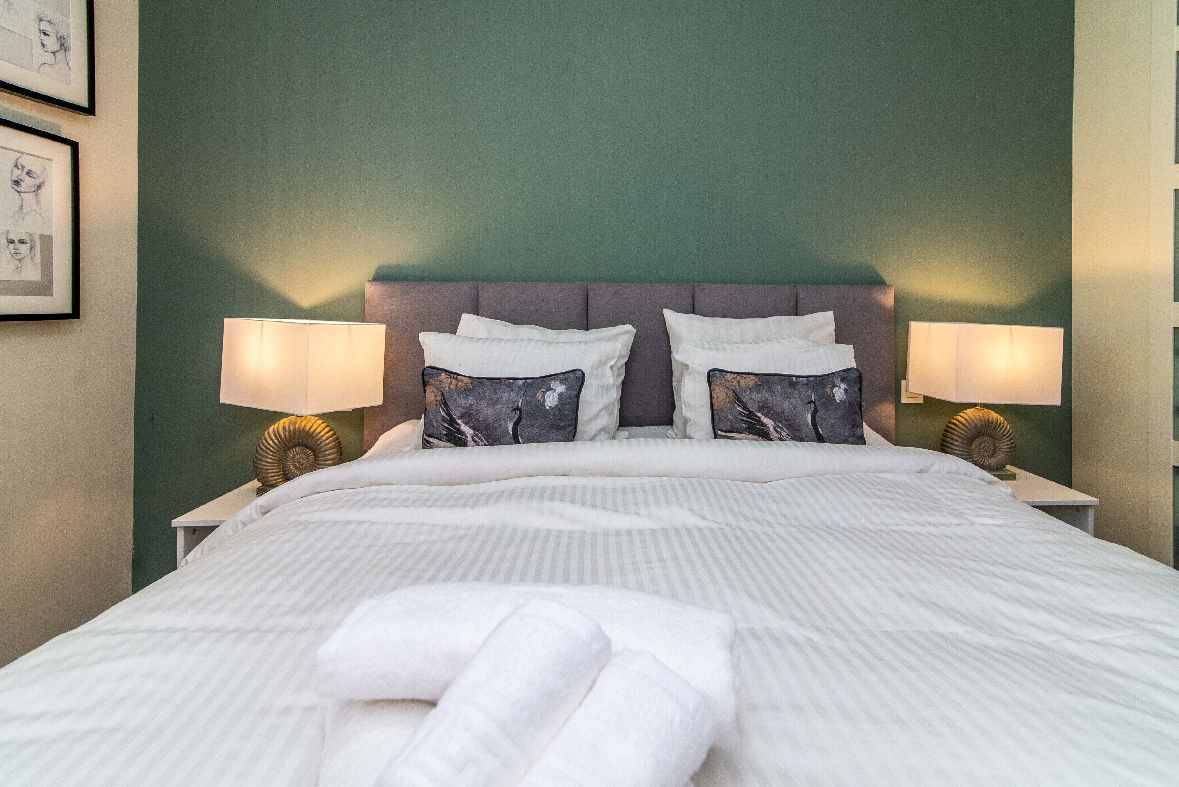 Una cama bien hecha con sábanas blancas y dos almohadas, cabecero, lámparas de noche y una pared con obras de arte enmarcadas.