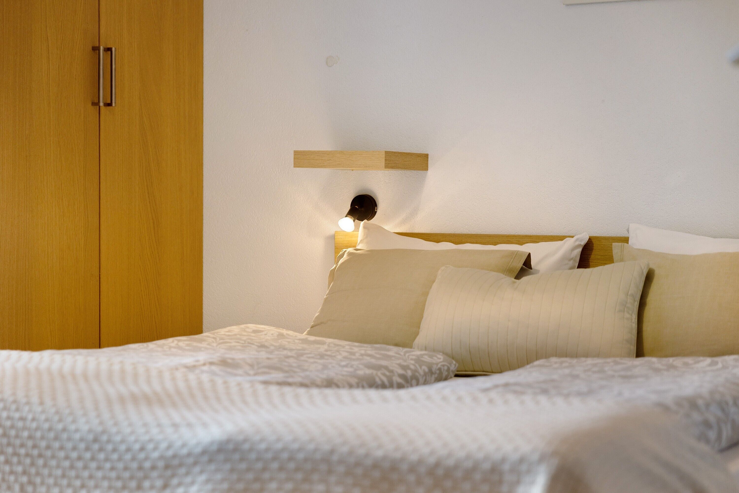 A bedroom with a wooden wardrobe, a bedside table with a lamp, and a bed with multiple pillows.