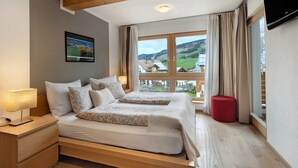 A bedroom with a large bed, a nightstand, a red ottoman, and a window with a view of a village.