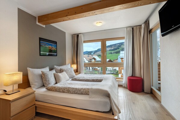 A bedroom with a large bed, a nightstand, a red ottoman, and a window with a view of a village.