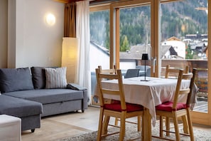 Una sala de estar moderna con un sofá gris, una mesa de comedor de madera con sillas, un ventanal con vistas a la montaña y una lámpara de pie.