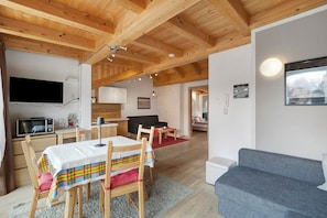 A cozy room with a dining table, chairs, a sofa, a TV, and a kitchen area.