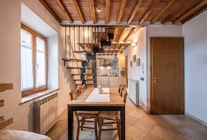 A dining area with a wooden table and chairs, a radiator, a window with curtains, a wooden door, and a kitchen in the background.
