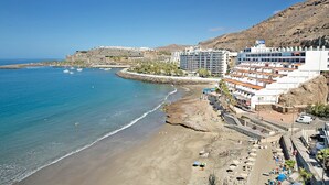 Ein Hotel an der Küste mit mehreren Etagen, ein Strand im Vordergrund und eine felsige Hügelsicht im Hintergrund.