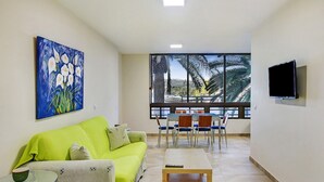 Ein Wohnzimmer mit einem grünen Sofa, einem Gemälde, einem Essbereich mit Stühlen und einem an der Wand befestigten Fernseher.
