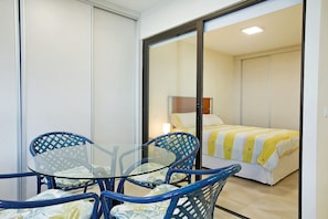Quarto com mesa de vidro e cadeiras azuis, cama com manta listrada e porta deslizante.