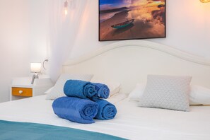 A neatly made bed with rolled blue towels, a white headboard, and a wall-mounted picture of a sunset over water.
