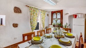 A dining area with a table set for four, chairs, a window with curtains, a refrigerator, and a wall with a rock and a hole. - Vacation Home 'Casa Bene' with Mountain View, Wi-Fi and Air Conditioning (Guimar)