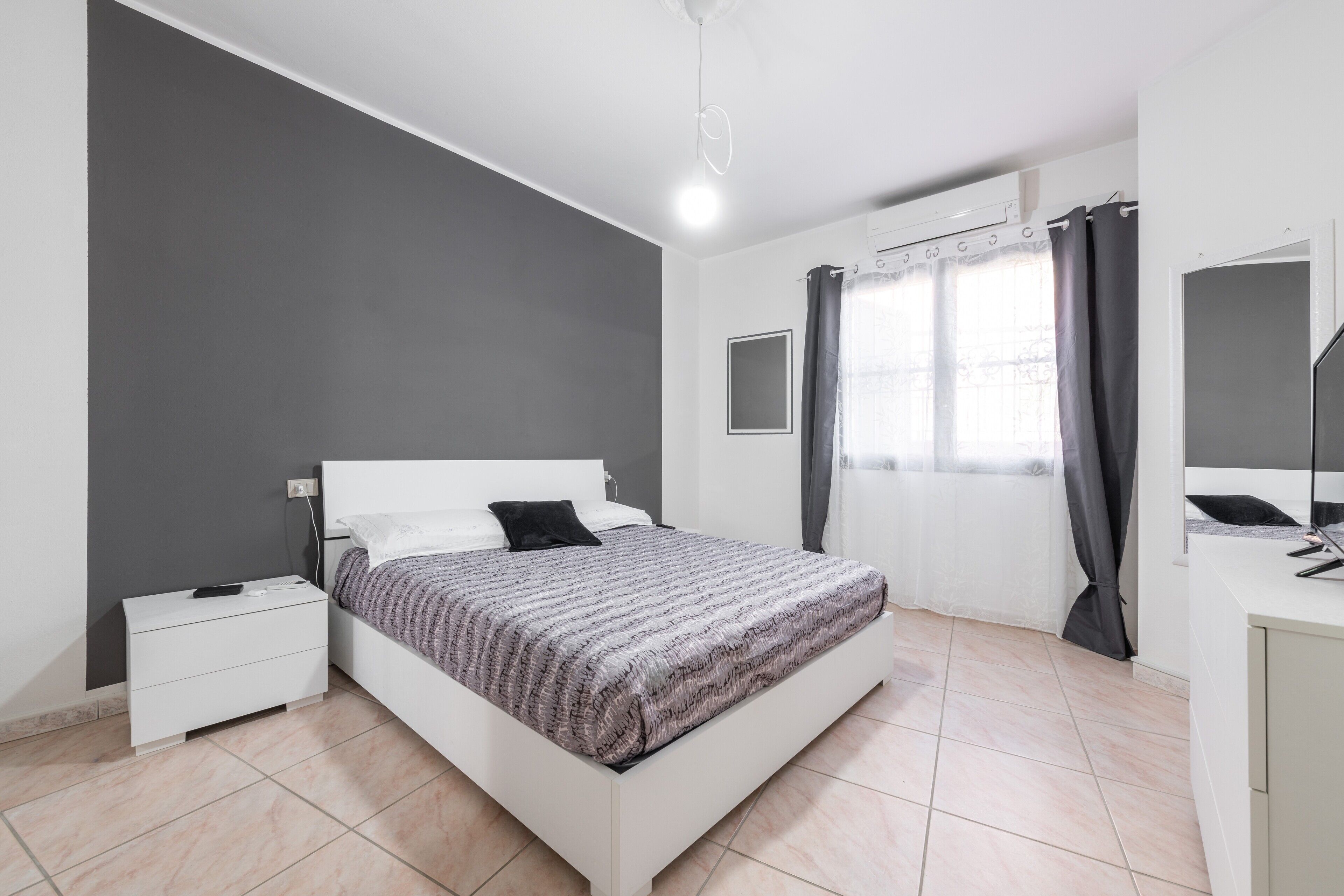 Ein Schlafzimmer mit einem Bett, einem Nachttisch, einer Kommode, einem Fenster mit Vorhängen und einer Deckenleuchte.