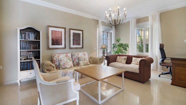Una sala de estar con sofá, sillón, mesa de centro, estantería, candelabro y vistas al exterior.