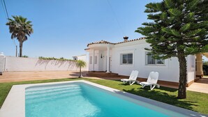Una piscina con dos sillas reclinables blancas, un edificio blanco con techo de tejas y una palmera.
