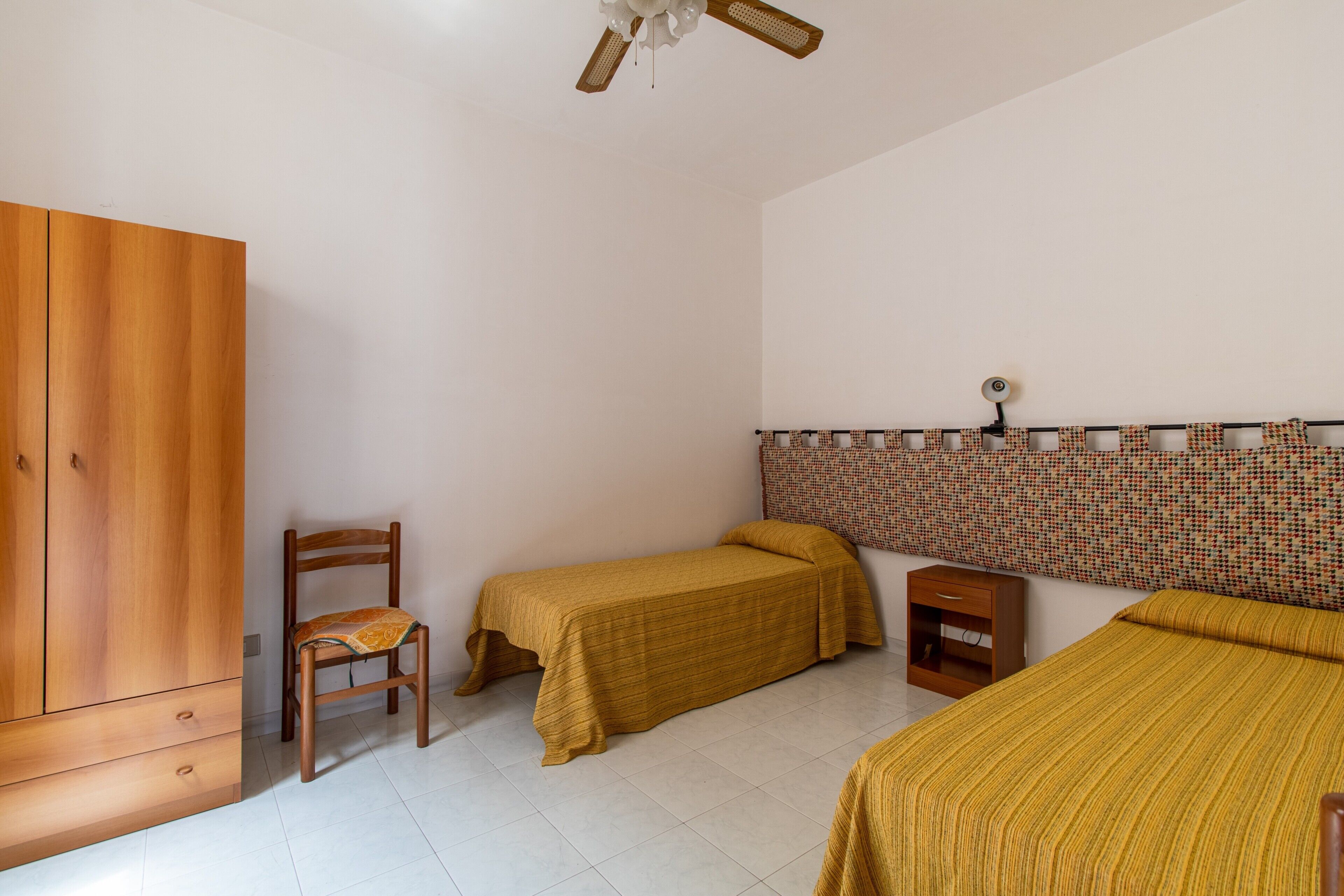 A bedroom with a wooden wardrobe, two single beds, a chair, a small bedside table, and a ceiling fan.