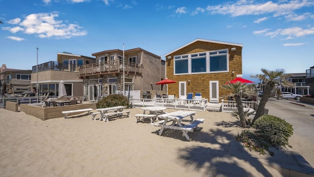 Une propriété située en bord de mer, comprenant un bâtiment à deux étages, un espace extérieur aménagé avec des sièges et une terrasse sablonneuse.