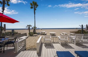 A beachside deck with wooden chairs and tables, a red umbrella, and a view of the beach and ocean.