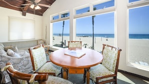 Une pièce avec un plafond en bois, une table ronde en bois entourée de chaises, de grandes fenêtres donnant sur la plage et une chaise de plage à l’extérieur.