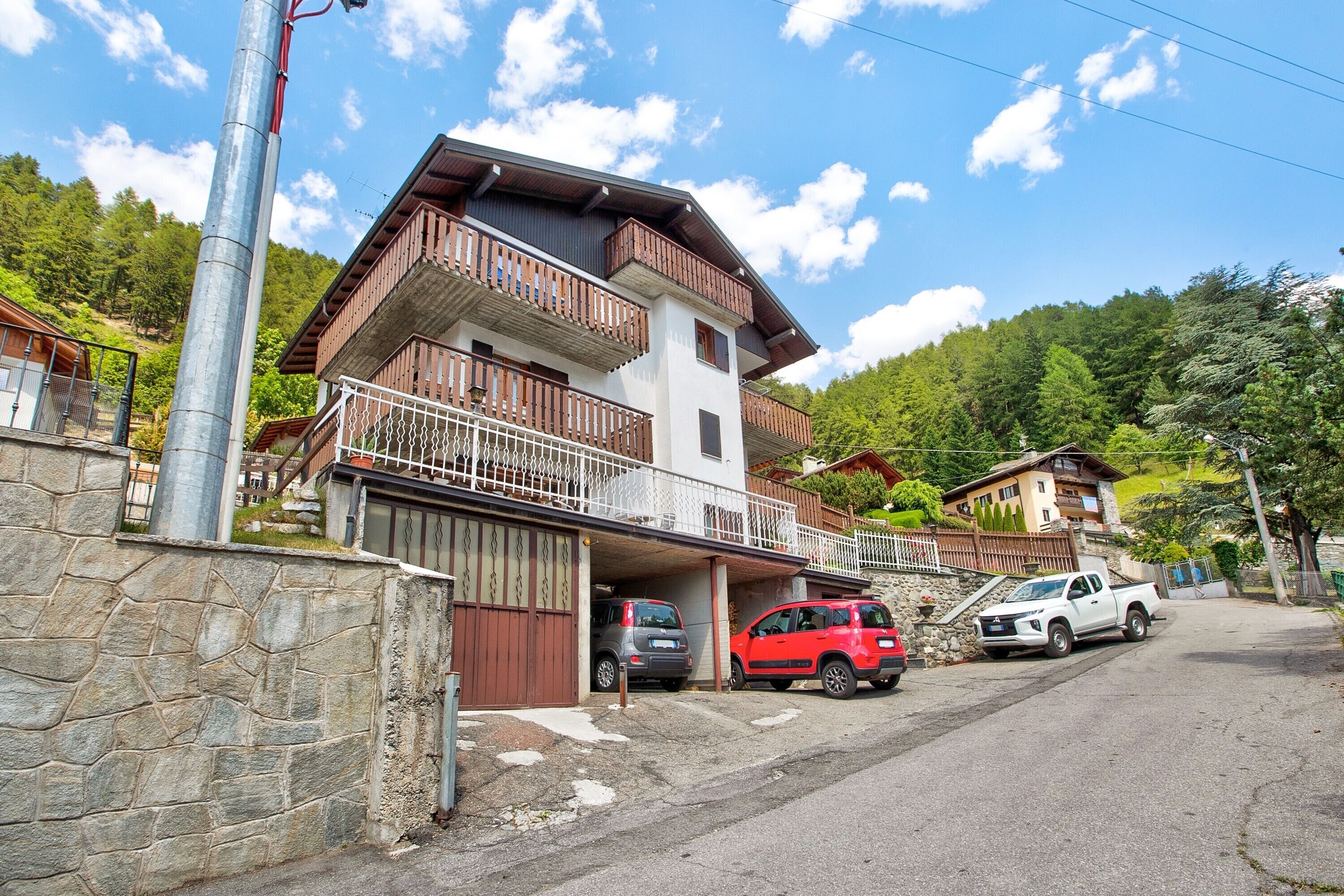 A two-story building with a balcony, stone wall, and parked vehicles.