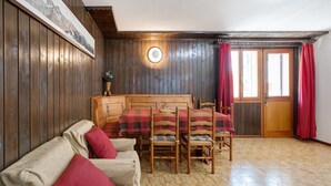 Ein Zimmer mit Holzwänden, einem Bett mit rot-schwarzem Karomuster, einem Holztisch mit Stühlen und einer runden Wanduhr.