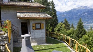 Un chalet de montagne rustique, doté d’une cheminée en pierre, d’une petite porte en bois et d’un balcon offrant une vue sur un paysage montagneux.
