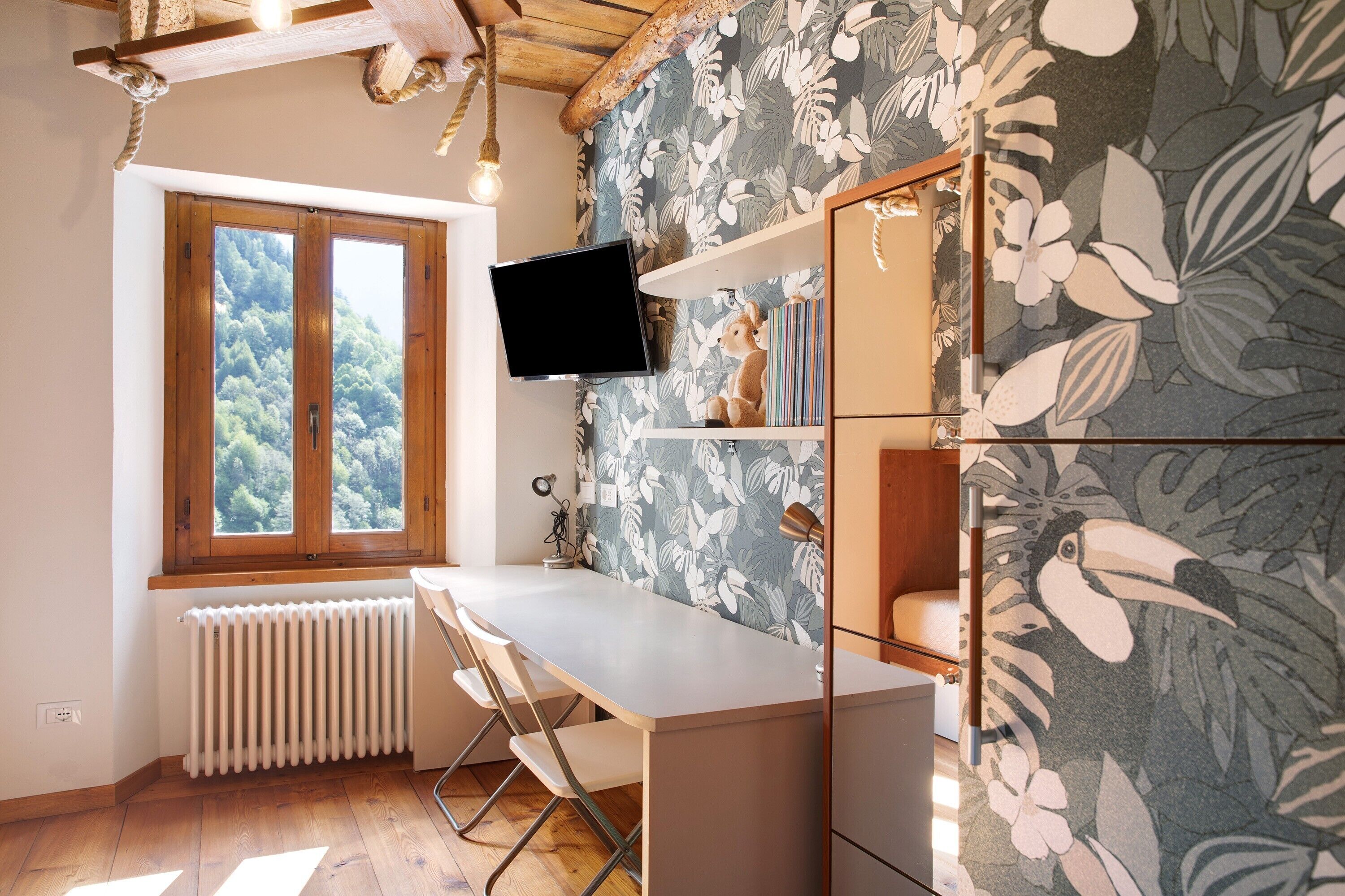 A room with a wooden desk, a radiator, a window with a view of trees, a mounted TV, and a wall with a floral pattern.