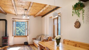 A cozy room with a wooden ceiling, a large window with a forest view, a wooden dining table, and a sofa. - Apartment 'Bellavista' with Mountain View & Garden (Chiesa In Valmalenco)