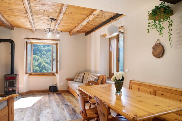 A cozy room with a wooden ceiling, a large window with a forest view, a wooden dining table, and a sofa.