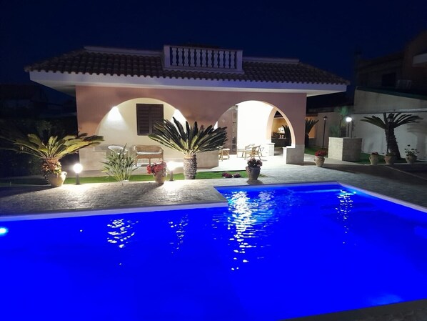 A swimming pool with a tiled deck, surrounded by potted plants and a building with arches.