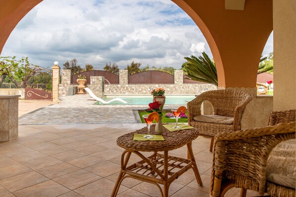 A covered outdoor seating area with wicker furniture, a pool, and a stone wall in the background.