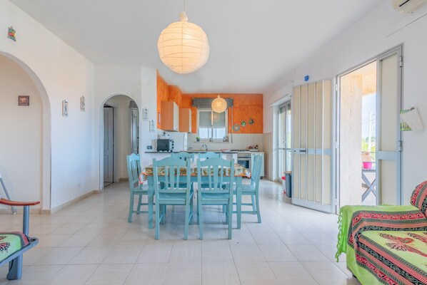 Amplia sala de estar con mesa de comedor, sillas y sofá. Se observa un área de cocina con armarios y una ventana.