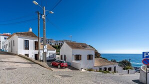 Una calle adoquinada en un pueblo costero con edificios blancos, un coche rojo estacionado al lado y un cartel de 'prohibido estacionar'.