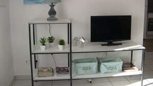 Una sala de estar con una televisión de pantalla plana, una lámpara y una estantería con libros y objetos decorativos.