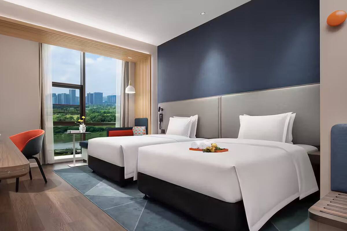 A hotel room with two beds, a desk, a chair, a TV, and a view of the city.