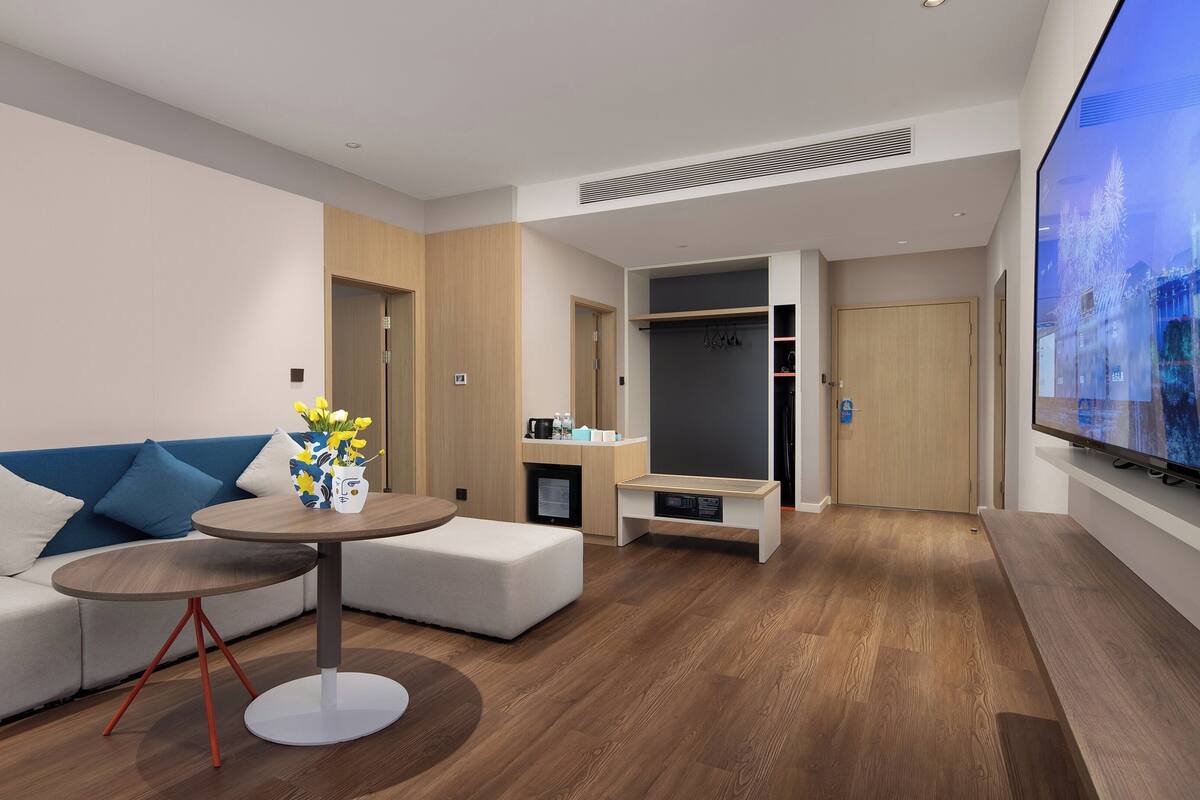 A modern hotel room with a sofa, a round table with a vase of flowers, a television, a wardrobe, and a small seating area.