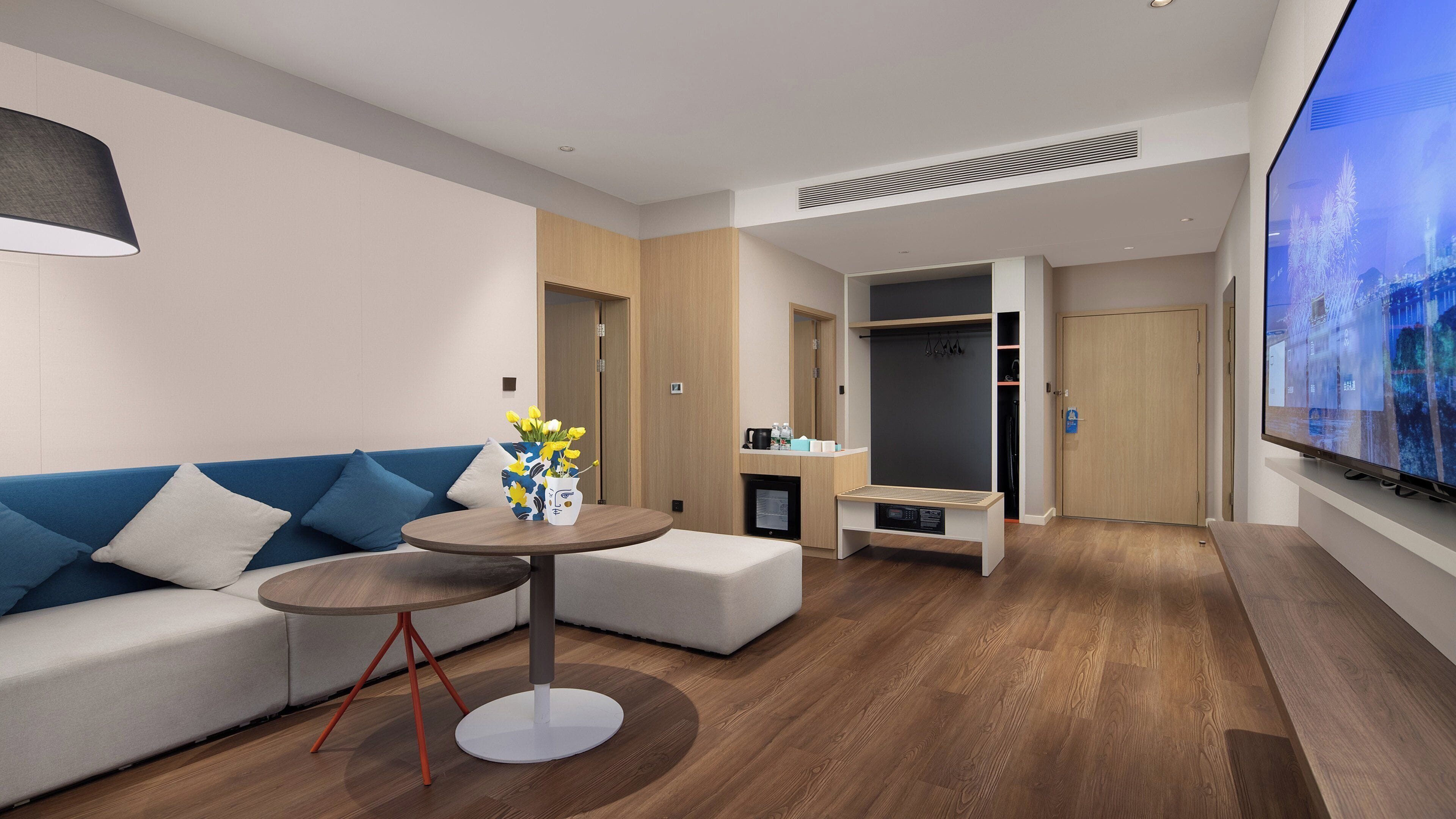 A modern hotel room with a sofa, a round table with a vase of flowers, a television, a wardrobe, and a small seating area.